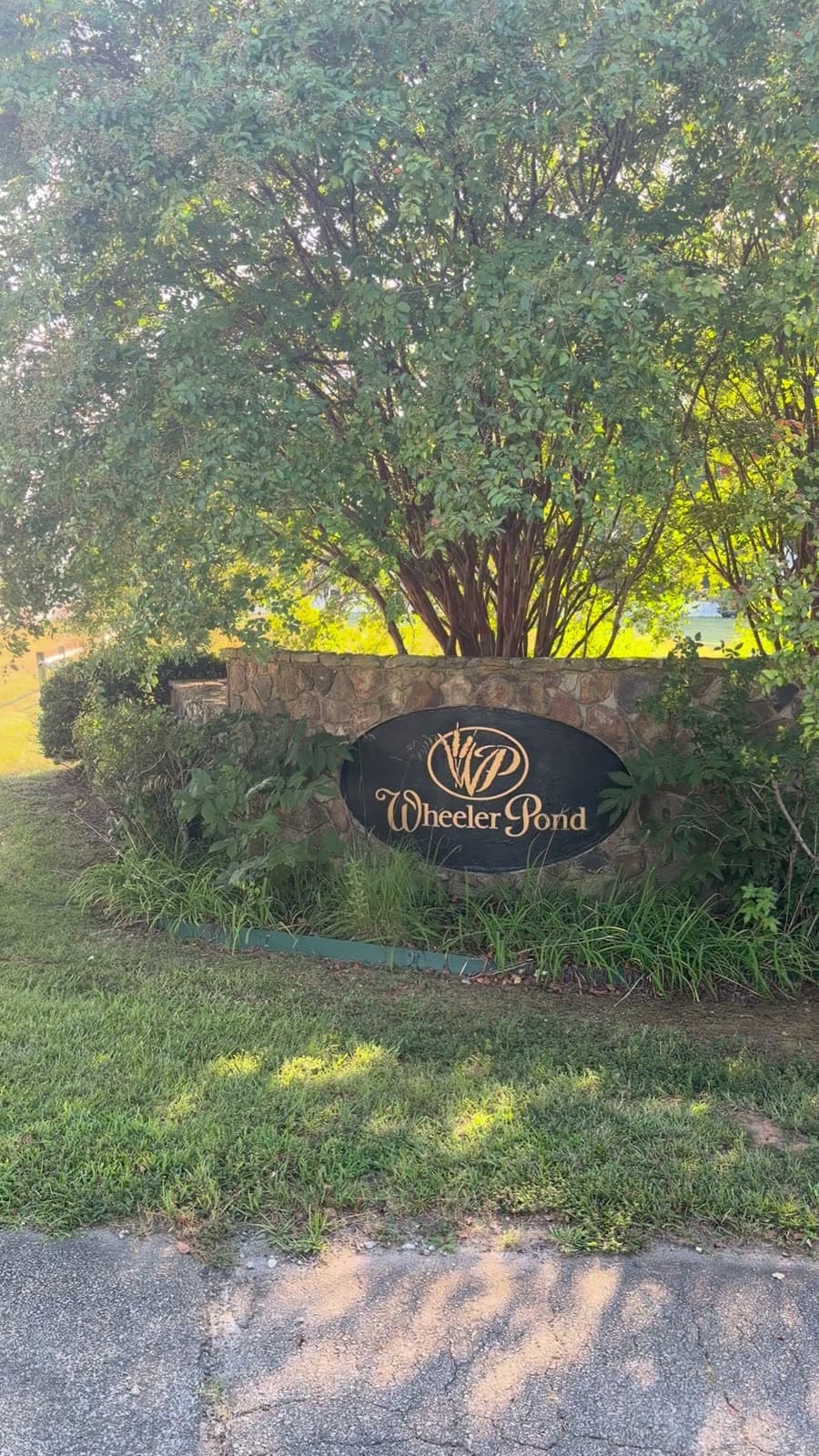 The stone entrance wall, carrying a black oval sign lettered Wheeler Pond in gold script above a WP monogram, with a crape myrtle growing behind it.